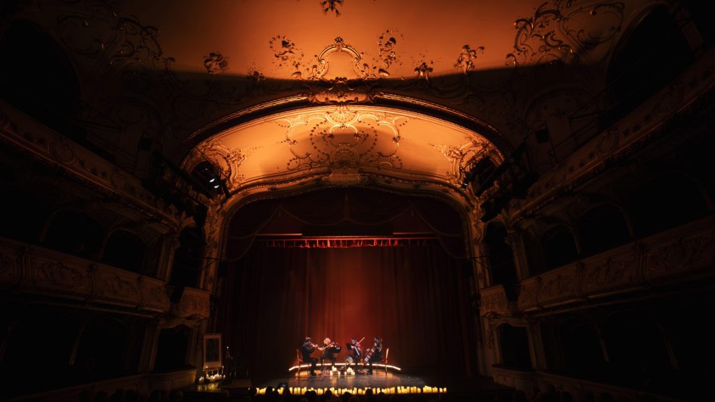 Ornate historic theater with a gilded stage; string quartet performing under warm amber lights.